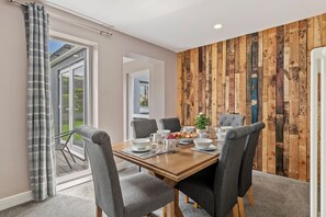 A dining area with a wooden table set for six, grey upholstered chairs, and a view through a glass door to a balcony. - Roslen - Lovely Bungalow Near Beach and Village (Saundersfoot)