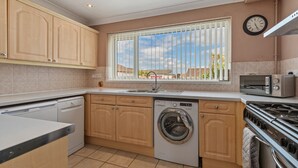 A modern kitchen with wooden cabinets, a washing machine, a stove, and a window with blinds. - Roslen - Lovely Bungalow Near Beach and Village (Saundersfoot)