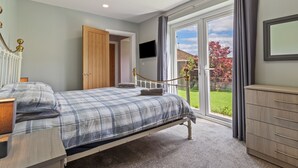 A bedroom with a bed, bedside table, dresser, TV, and a sliding glass door to a garden. - Roslen - Lovely Bungalow Near Beach and Village (Saundersfoot)