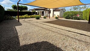 A covered outdoor area with a gravel ground, a wooden pergola, and a house in the background.
