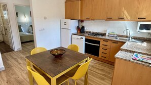 A compact kitchen with wooden cabinets, a dining table with yellow chairs, a refrigerator, a stove, and a dishwasher.