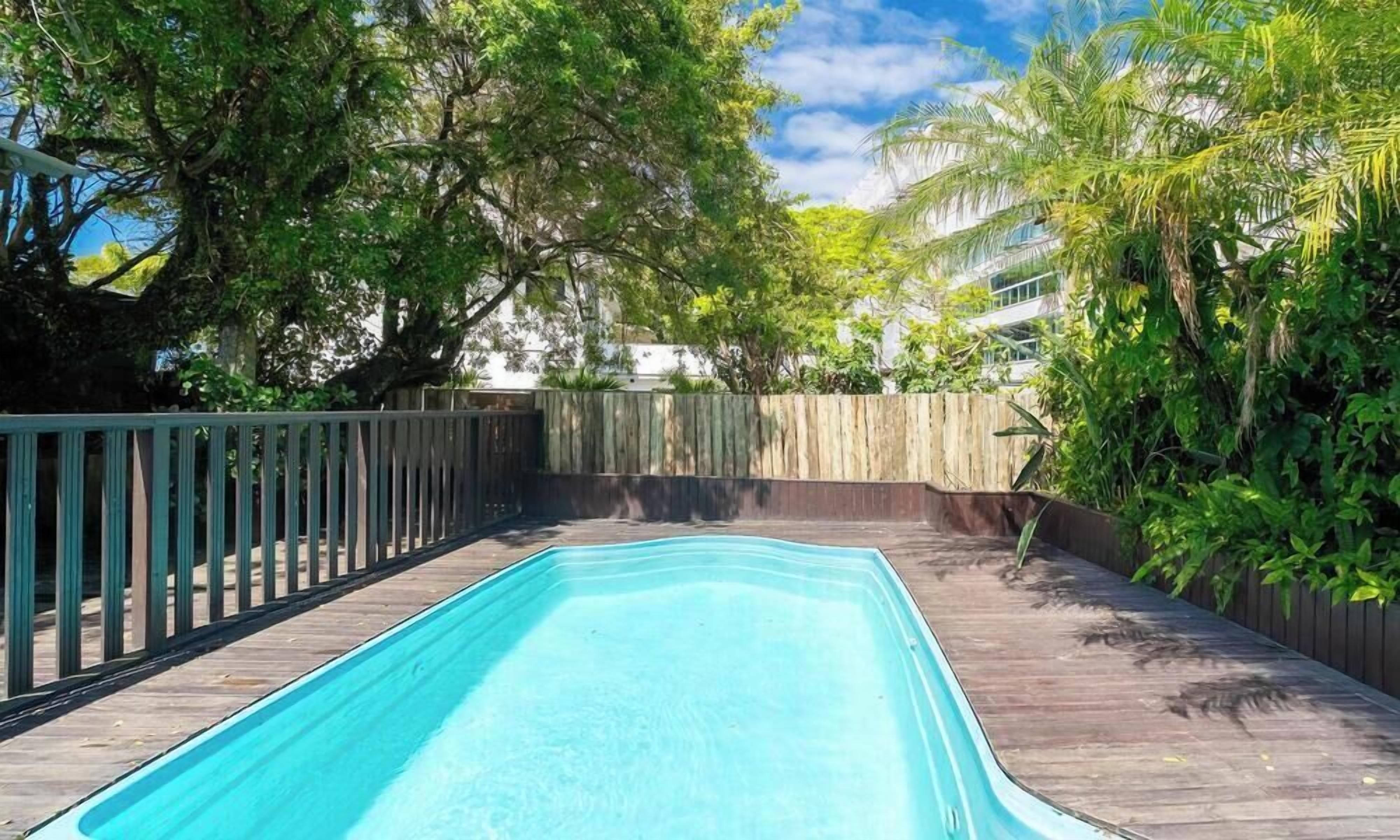 Une piscine rectangulaire entourée d’une terrasse en bois, le tout délimité par une clôture en bois et bordé d’une végétation luxuriante.