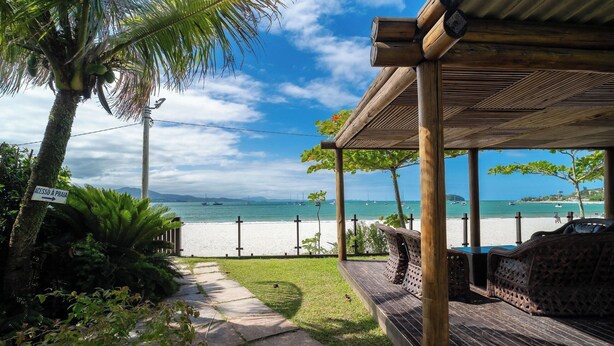 A beach view with a wooden pergola, a pathway, and a sign indicating access to the beach. - JBM Apto pé na areia com vista mar (Florianópolis)