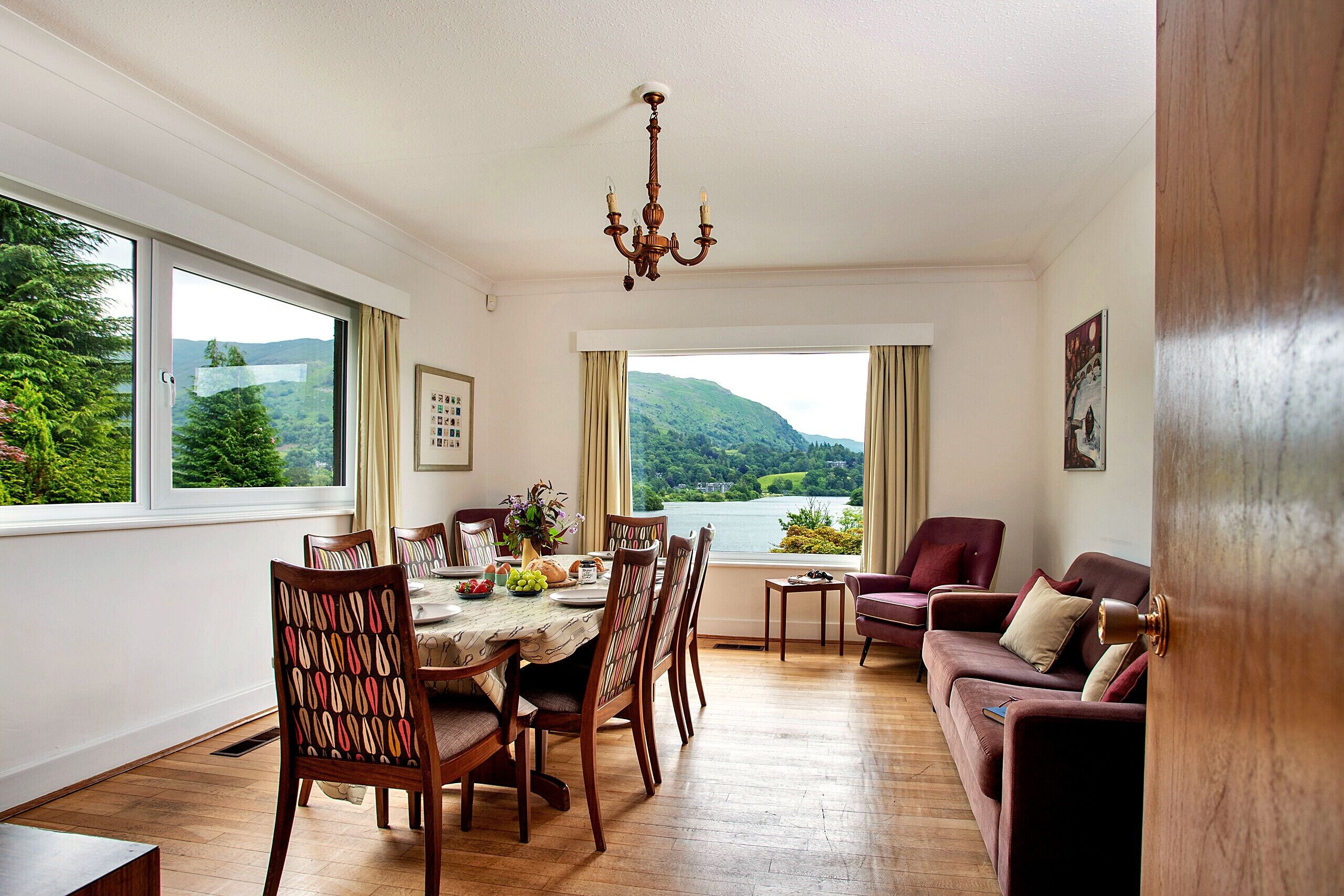 Un espace repas avec une table dressée pour six personnes, un lustre et une vue sur un lac et des collines, visible à travers de grandes fenêtres.