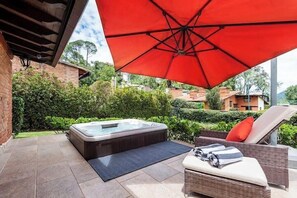A hot tub under a red umbrella, a wicker lounge chair, and a tiled patio area.