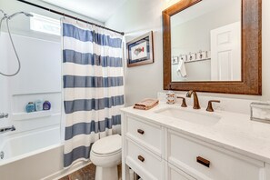 Un baño con bañera, cortina de ducha, inodoro, lavamanos con espejo y un cuadro en la pared.