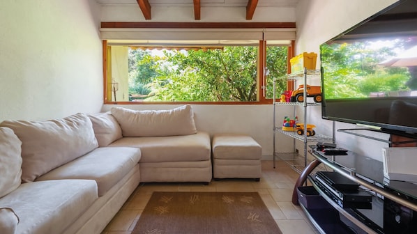 A living room with a beige sectional sofa, a television on a stand, and a window with a view of greenery. - Casa Male (Valle de Bravo)