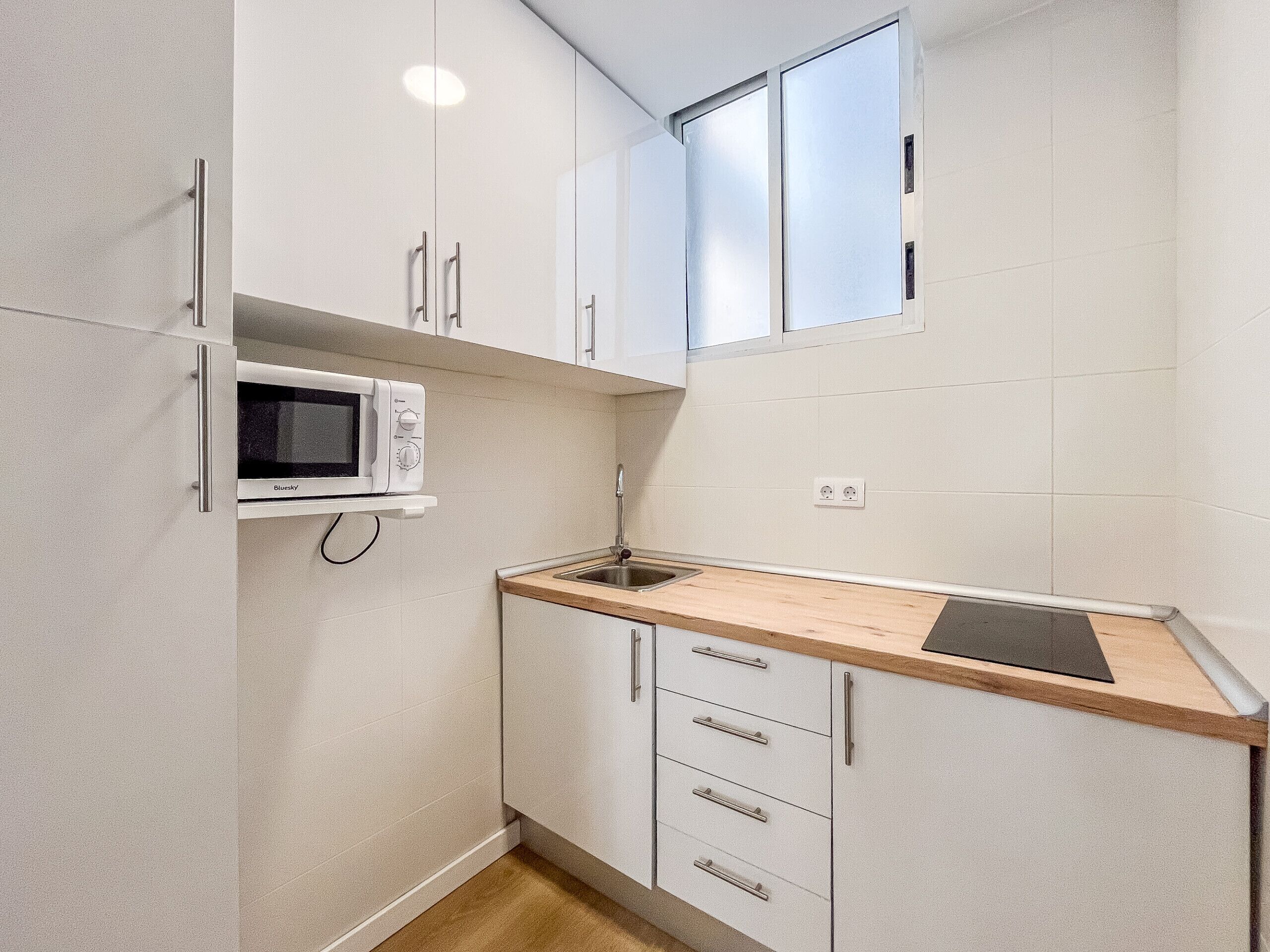 Cocina compacta con armarios blancos, microondas, pileta y ventana sobre la encimera.