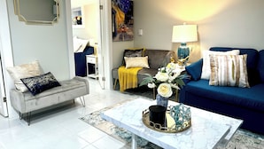Ein modernes Wohnzimmer mit einem grauen Sofa, einem blauen Sofa, einem weißen Couchtisch und einem grauen Sessel. An der Wand hängt ein Gemälde, und auf dem Couchtisch steht eine Vase mit Blumen. Eine Lampe ist ebenfalls vorhanden.