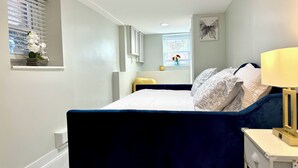 Ein Schlafzimmer mit Bett, Nachttisch, Lampe, Fenster mit Jalousien und einer kleinen Ablage mit Blumen.