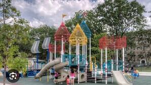 Ein Spielplatz mit verschiedenen Spielgeräten, darunter Rutschen und Kletterbereichen, umgeben von Bäumen und einem Gebäude im Hintergrund.