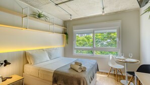 Ein modernes Schlafzimmer mit Bett, Nachttisch, Lampe und Fenster mit Blick auf Bäume.