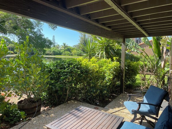 A covered patio area with a wooden table and chairs, surrounded by lush greenery and a view of a garden and a pond.