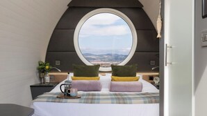 Un dormitorio con un amplio ventanal circular, una cama con ropa de cama escocesa, dos almohadas, una mesita de noche con lámpara y vistas al paisaje.