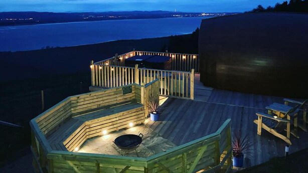 Una terraza de madera con un área de estar circular y un brasero, con vista a un cuerpo de agua y luces de la ciudad a lo lejos.