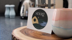 Tarjeta con el logo de 'The Snug at Logie Farm', una cafetera y una taza de cerámica sobre una superficie de madera.