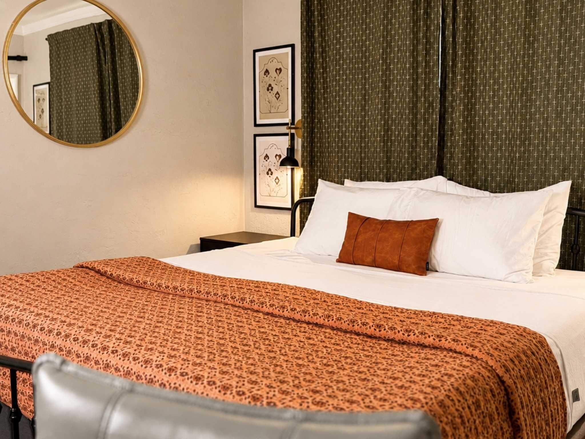 Une chambre d’hôtel avec un lit, un miroir rond, des œuvres d’art encadrées et un rideau à motifs.