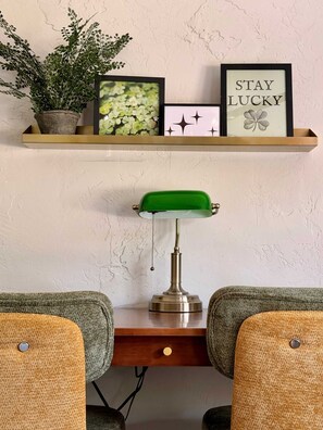 Un bureau avec une lampe de bureau verte, deux chaises, une étagère avec une plante et des tableaux encadrés, dont l’un porte l’inscription « QUE LA CHANCE SOIT AVEC VOUS ».