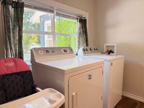 A laundry room with a front-loading washer and dryer, a small shelf with a water heater, and a window with curtains.