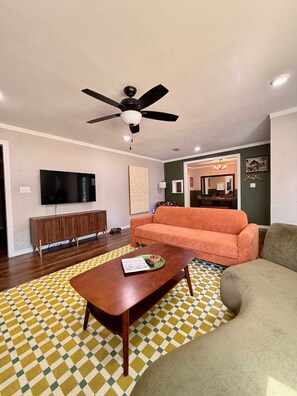 Un salon doté d’un ventilateur de plafond, d’un téléviseur à écran plat, d’un meuble en bois, d’un canapé et d’un tapis à motifs.