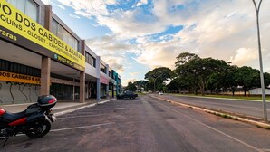 A street view with a motorcycle parked on the side, a building with a sign for 'Quins - Closet - Expositores Em Geral', and a clear sky with clouds.
