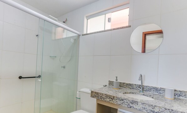 A bathroom with a glass shower enclosure, a sink with a granite countertop, and a small window above the mirror. - PPRAs Casas ao lado da praia Barramares (Ilhéus)