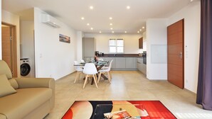 A modern living room with a sofa, a dining area with a table and chairs, a kitchen with cabinets and a window, and a hallway with a door.