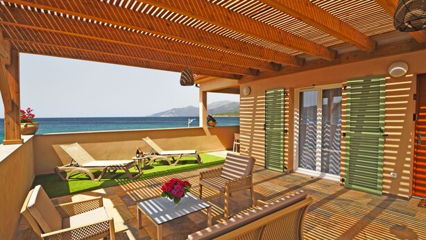 A sunlit outdoor seating area with a view of the sea, wooden furniture, and a table with a vase of flowers.