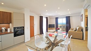 A modern apartment with a glass dining table, white chairs, a kitchen with a stove, and a view of the sea.