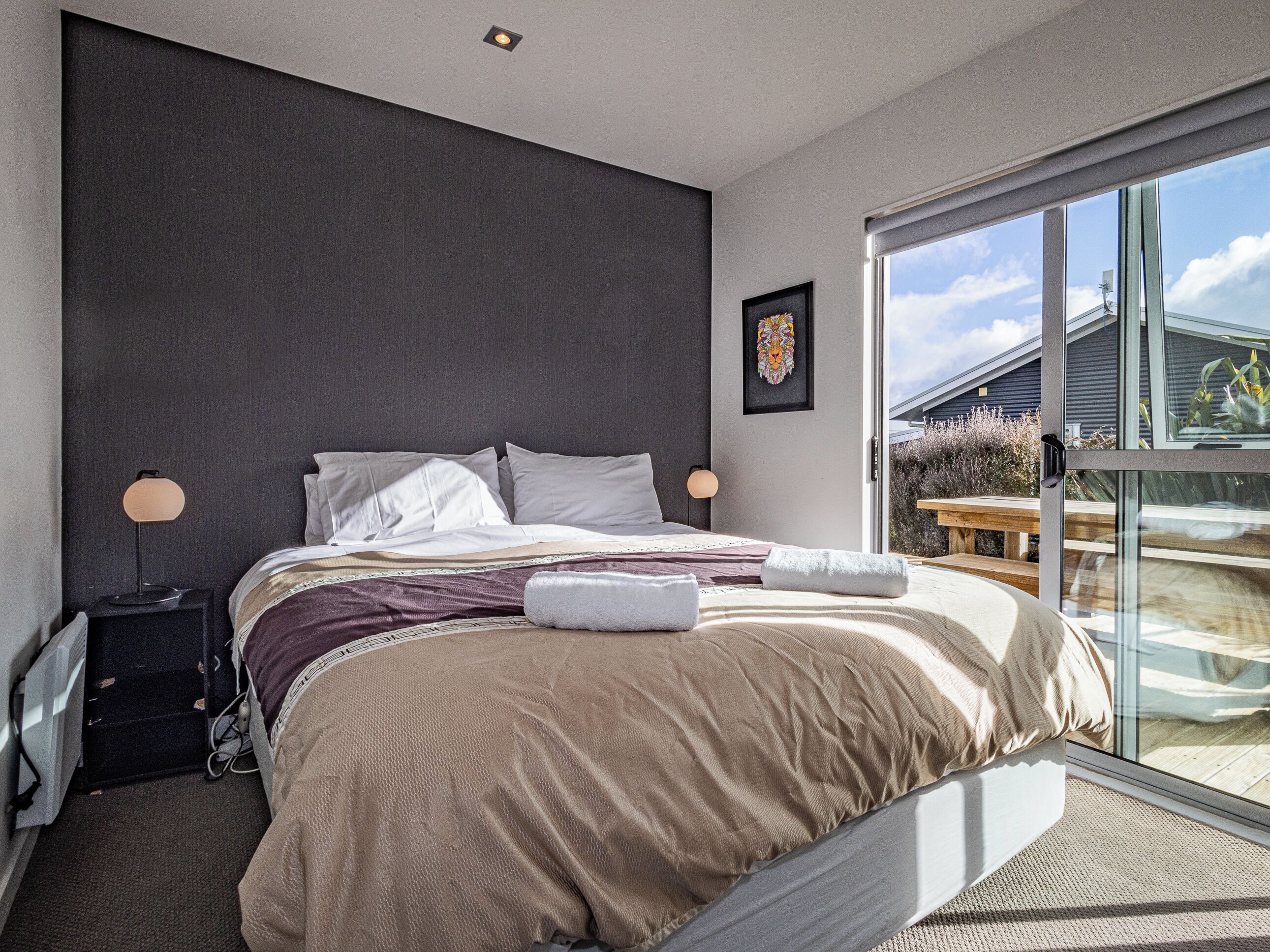 A modern bedroom with a large bed, a bedside table, a lamp, and a view of the outdoors through a sliding glass door.