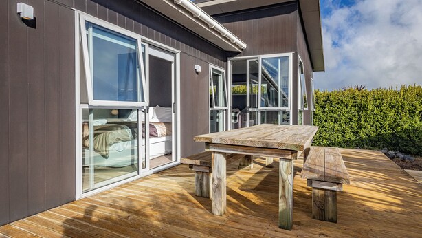 Une maison moderne avec de grandes portes vitrées donnant sur une chambre, une terrasse en bois et un ciel dégagé.