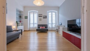 A spacious living room with a grey sofa, a red cabinet, and a TV. There are two windows with curtains, a bookshelf, and a hanging light fixture. - Appartamento Re Umberto by Wonderful Italy (Turin)
