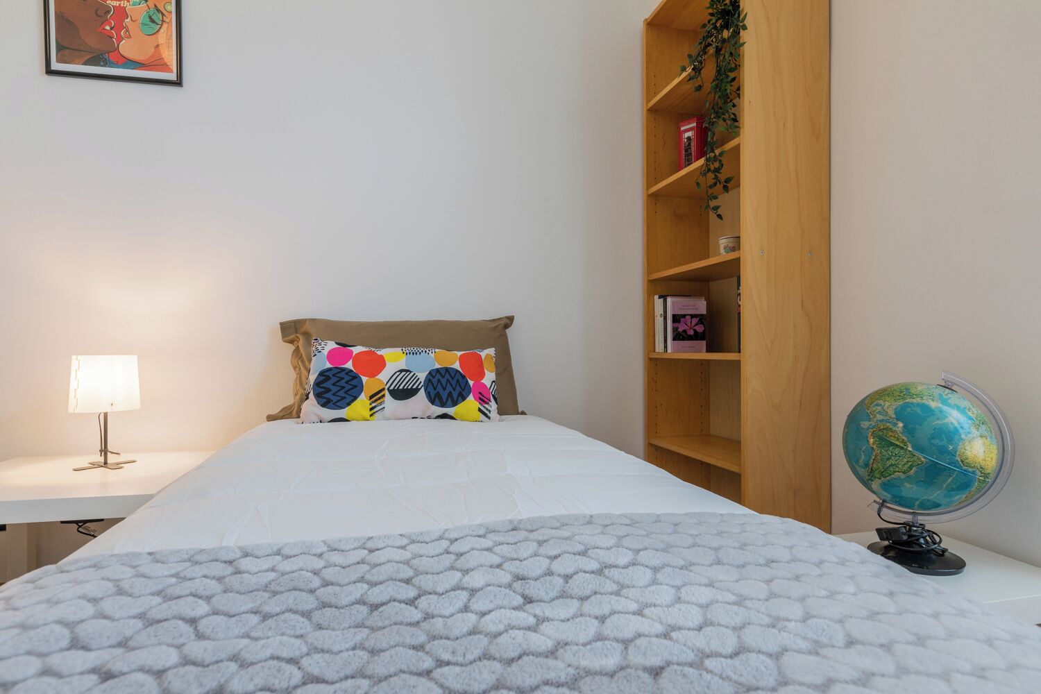 Une chambre à coucher bien rangée, avec un lit, une lampe de chevet, une bibliothèque remplie de livres et un globe terrestre, ainsi qu’un tableau encadré accroché au mur.