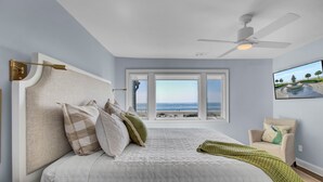 Une chambre avec un grand lit, un ventilateur de plafond, une fenêtre donnant sur la plage et un fauteuil avec un coussin.