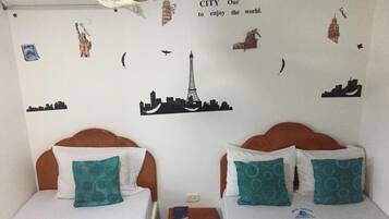A hotel room with two beds, each with a wooden headboard and teal pillows. The walls are decorated with cityscape decals and a ceiling fan.