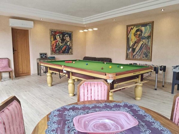 A pool table in a room with a wooden floor, a table with a patterned tablecloth, and two paintings on the walls.
