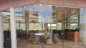 A glass-enclosed dining area with a round table and wicker chairs, a kitchen with wooden cabinets, and a view of the outdoors. - Domaine lamrah (Oulad Hassoune)