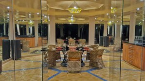 A spacious dining area with a high ceiling, multiple chandeliers, and a central round table surrounded by wicker chairs. - Domaine lamrah (Oulad Hassoune)