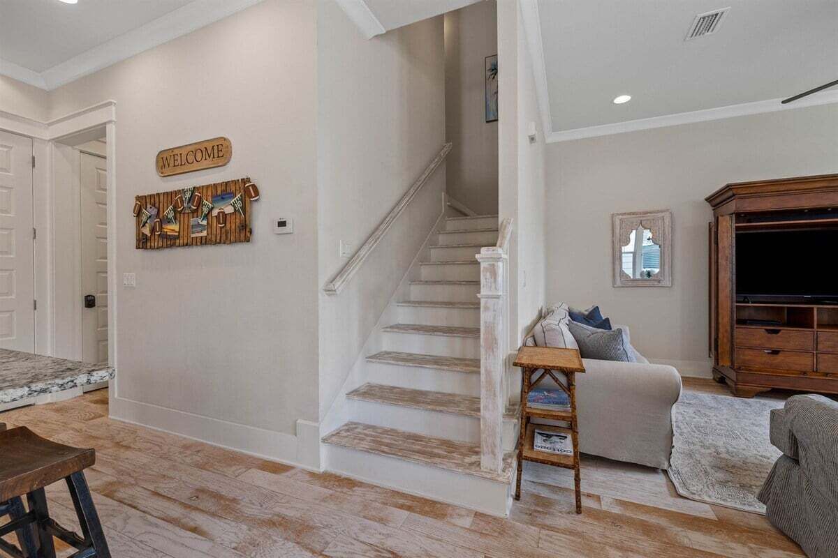 Un interior de hogar con una escalera, un cartel que dice 'WELCOME', un tablón de anuncios de madera, un sofá, un mueble para televisor y una mesa de comedor.