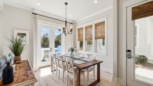 A dining area with a wooden table and white chairs, a chandelier, and a view through large windows with blinds. - Heated Pool - Golf Cart+Bikes - Close to Beach! (Inlet Beach)