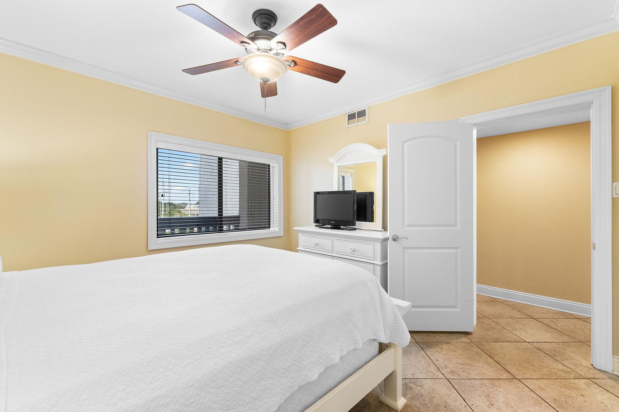Um quarto com uma cama, um ventilador de teto, uma televisão sobre um penteadeira e uma janela com persianas.