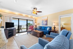 A spacious living room with a large TV, comfortable seating, a ceiling fan, and a balcony with a view. - Shipwatch A301 (Pensacola)