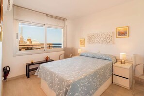 Un dormitorio con cama, mesitas de noche, un escritorio y ventana con vista a edificios y al mar.