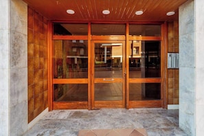 Entrada de un edificio con puerta de madera, paneles de vidrio, suelo de baldosas e iluminación empotrada en el techo.