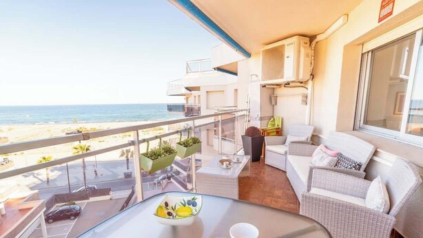 En balkon med udsigt over stranden, med fletmøbler og et bord med en skål frugt samt glas.