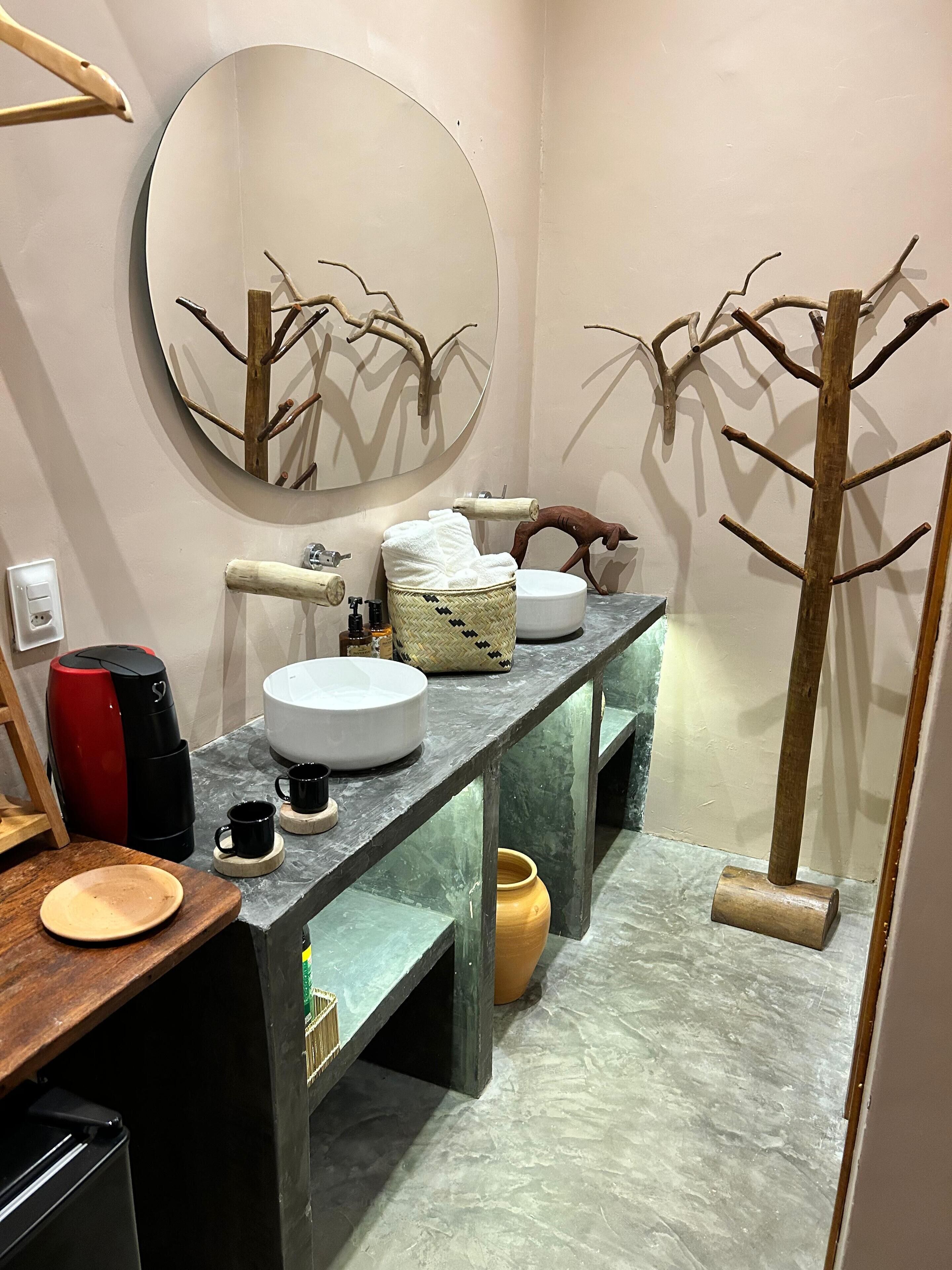 A modern bathroom with a round mirror, wooden branch decor, a sink, a coffee maker, and a towel rack.