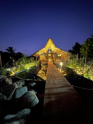 A thatched-roof structure with a paved pathway leading to it, surrounded by rocks and greenery.