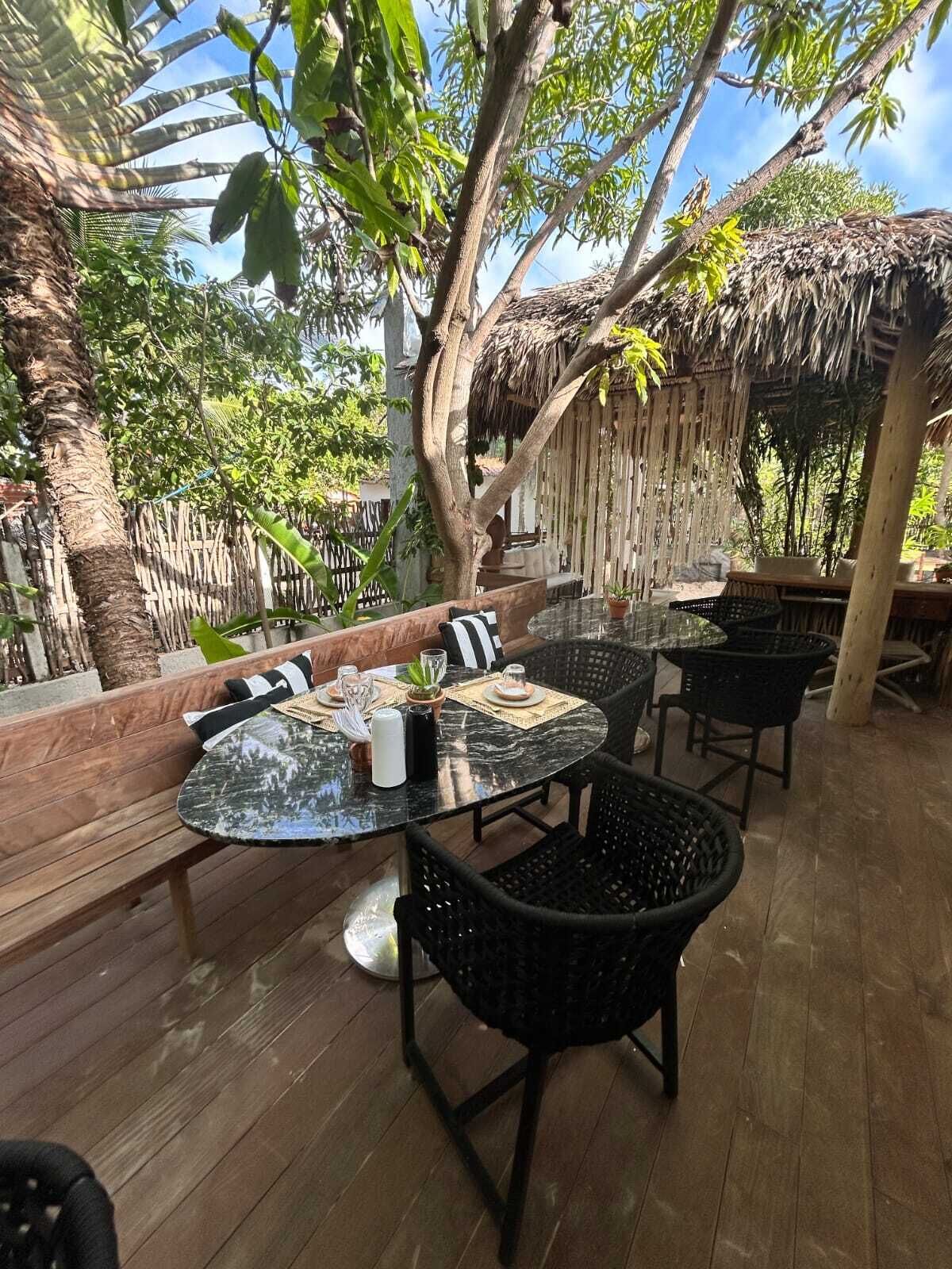 Un patio exterior con una mesa redonda de vidrio, sillas de mimbre negro y un banco de madera.