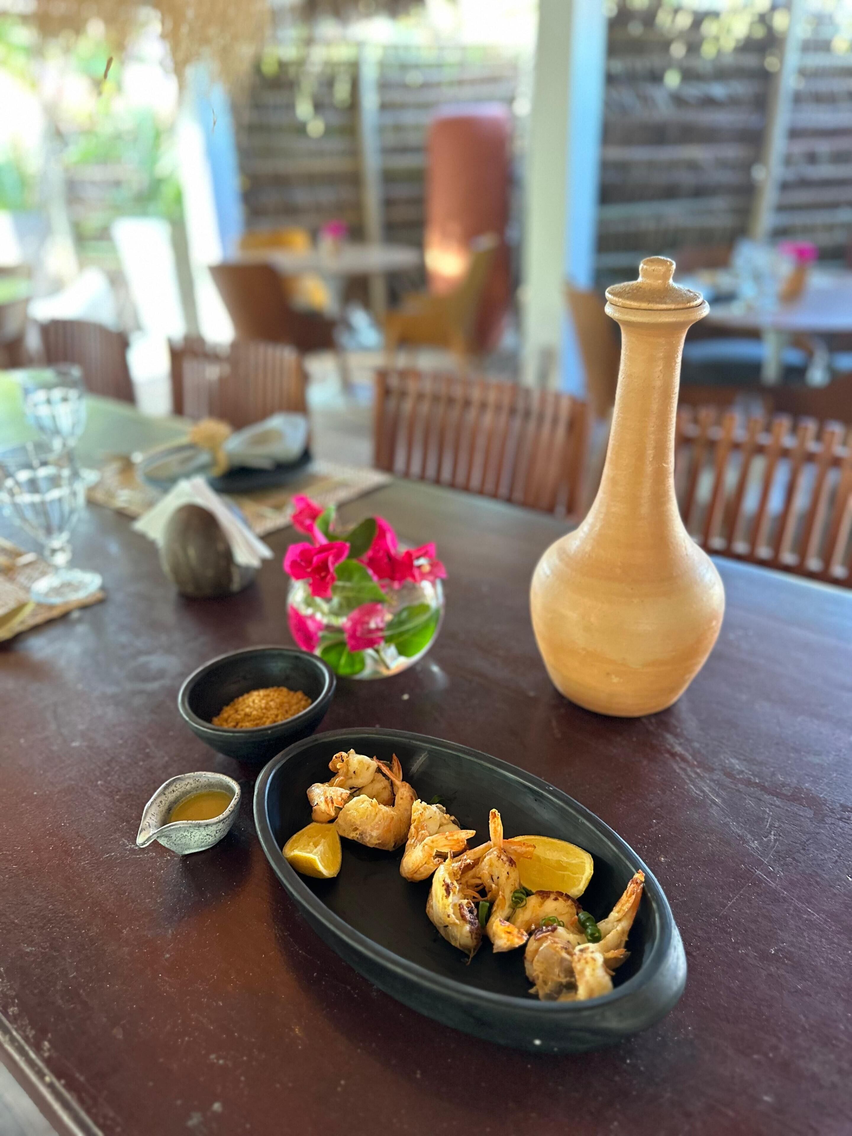 A dining table with a plate of grilled shrimp, a small bowl of sauce, and a decorative vase with flowers.
