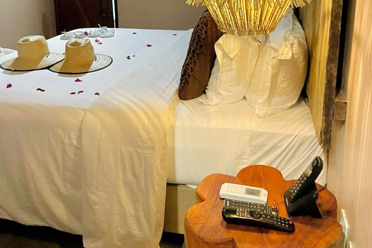 A hotel room with a bed, a hanging lamp, a wooden side table with a remote control, and a door in the background.